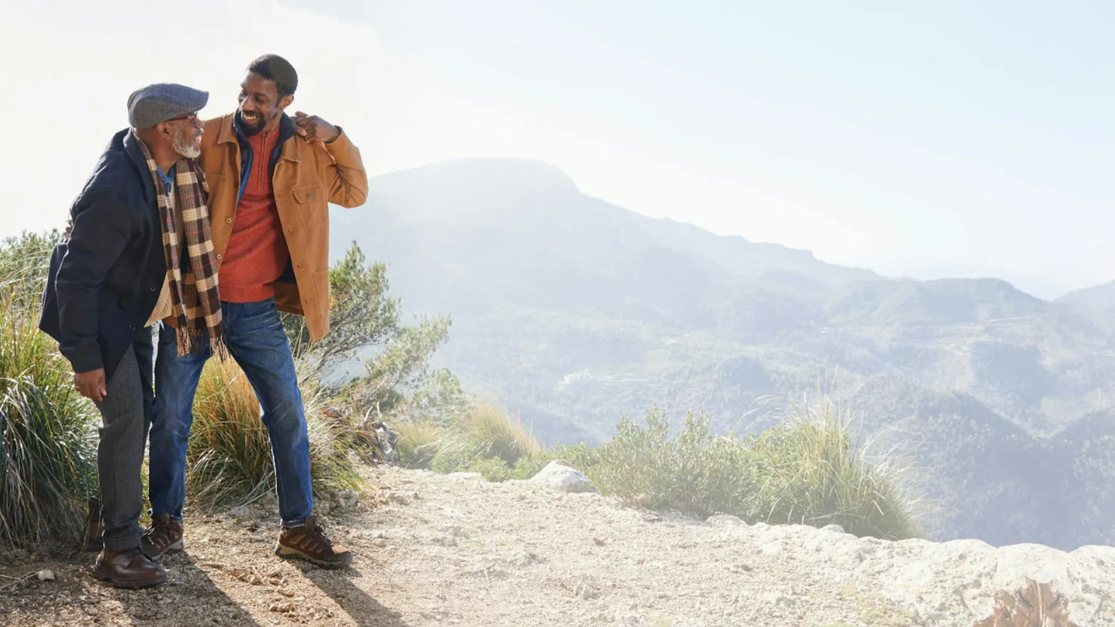 Two men in jackets laughing together on a mountain trail above a wide valley