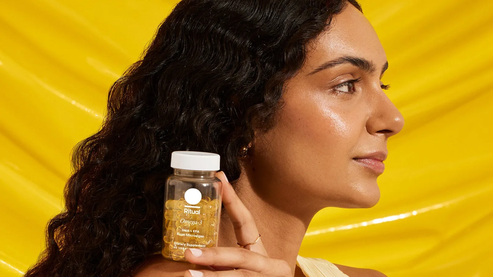 A woman in profile holding a bottle of Ritual Omega-3 capsules at her shoulder against a bright yellow backdrop