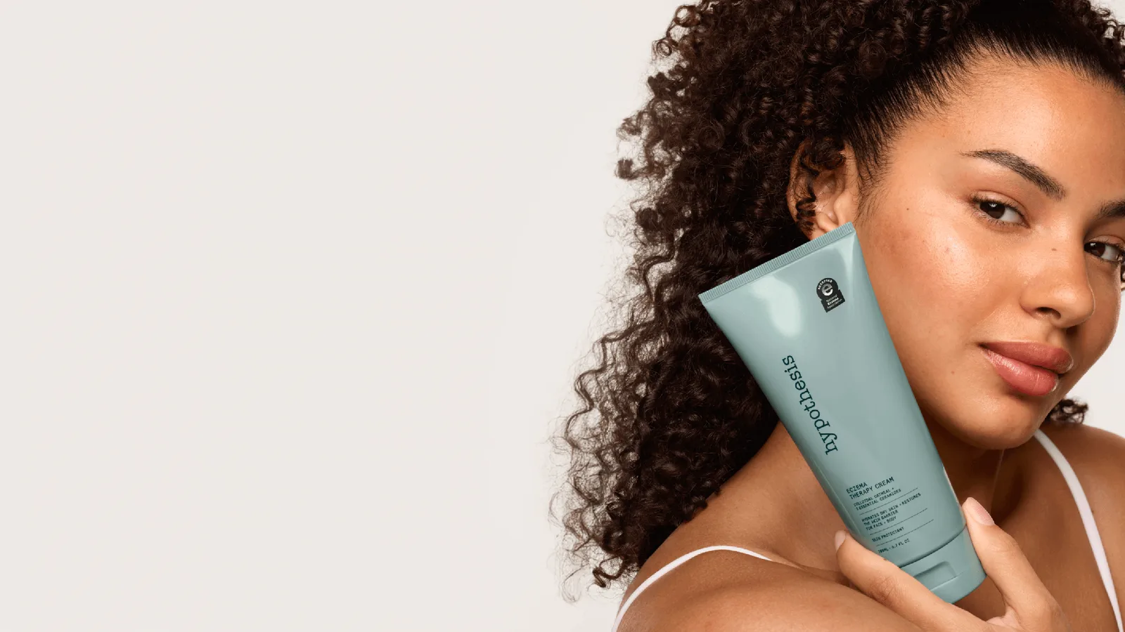 A woman with curly hair holding a tube of Hypothesis Eczema Therapy Cream against her cheek
