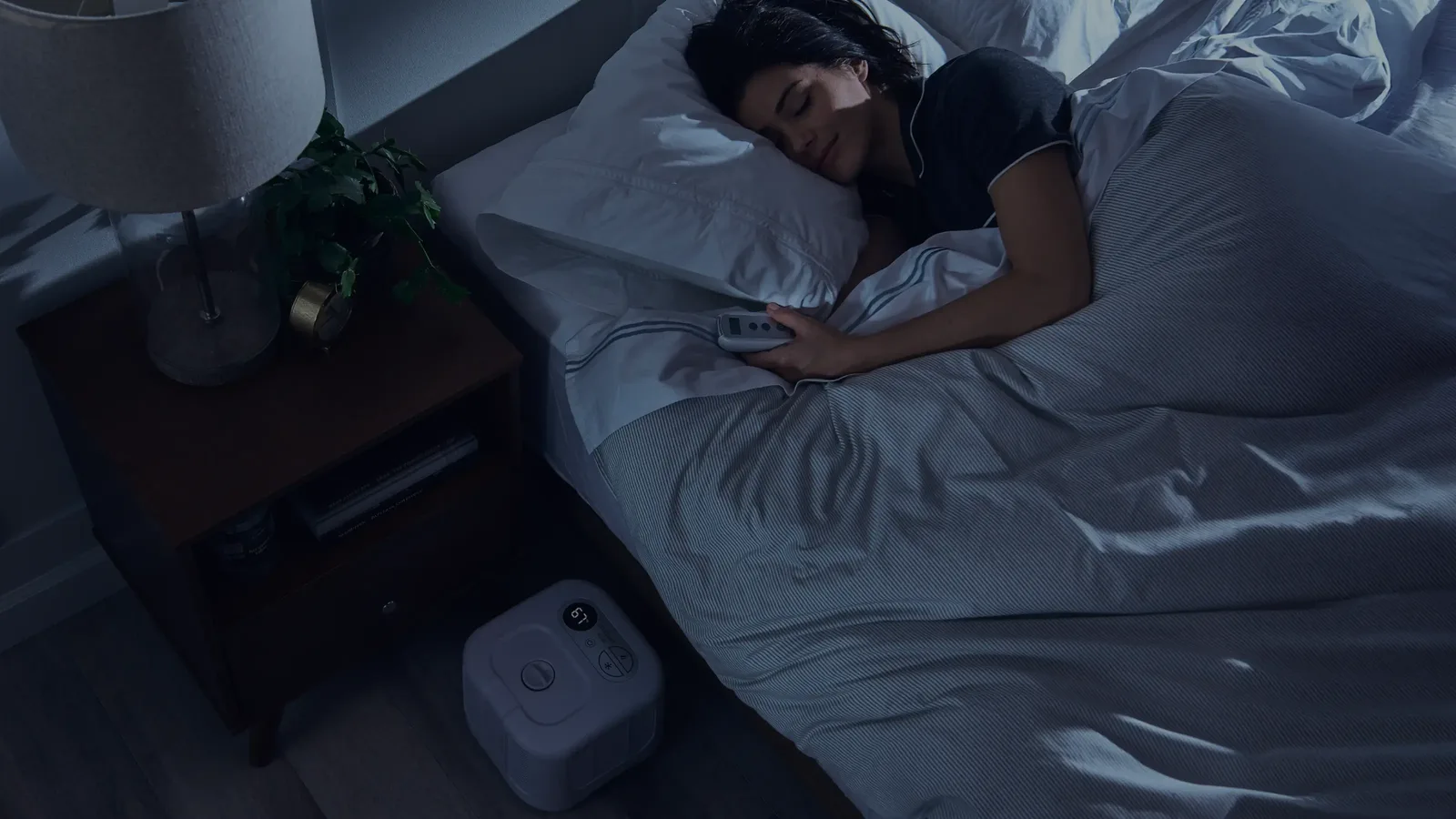 A woman asleep on her side in a dim bedroom, holding a remote, with a Chilipad Cube bed cooling unit on the floor beside the bed