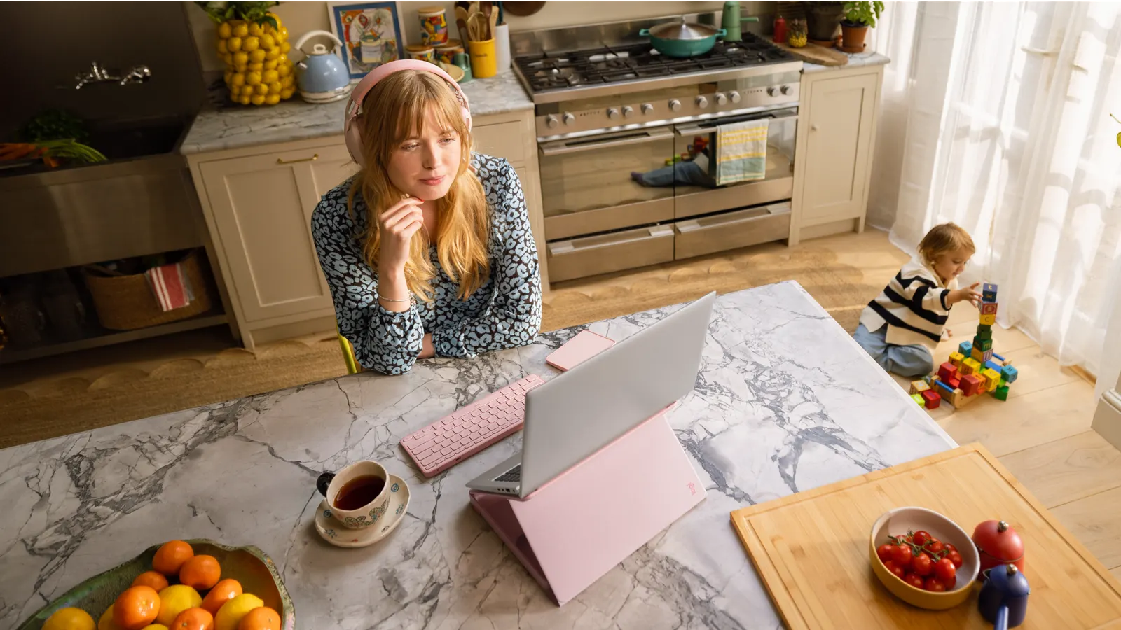 A woman working at a kitchen island with her laptop raised on a stand, a toddler playing with blocks nearby
