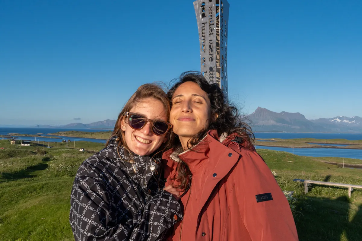 Two friends smiling with eyes closed in bright sunlight, fjord and mountains in the background